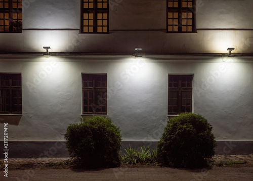White building wall illuminated by night lamps with bushes