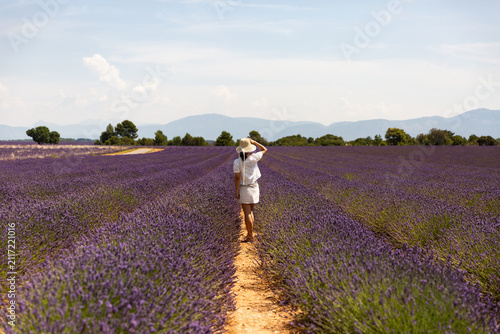 Une femme habillée en blanc debout au milieu d'un champ de lavande violette en fleur, elle porte un chapeau et regarde l'horizon, arbres verts, collines et ciel bleu en arrière-plan, sud de la France
