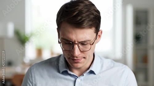 A young professional man wearing optical eyewear focuses intensely while looking down at his digital work inside a bright home office environment during his daily productive remote business