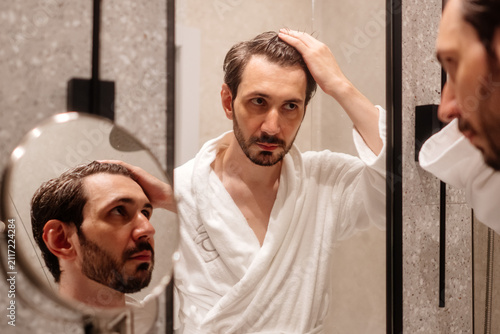Quadro su tela Man in a white robe examining his hair and receding hairline in a bathroom mirro
