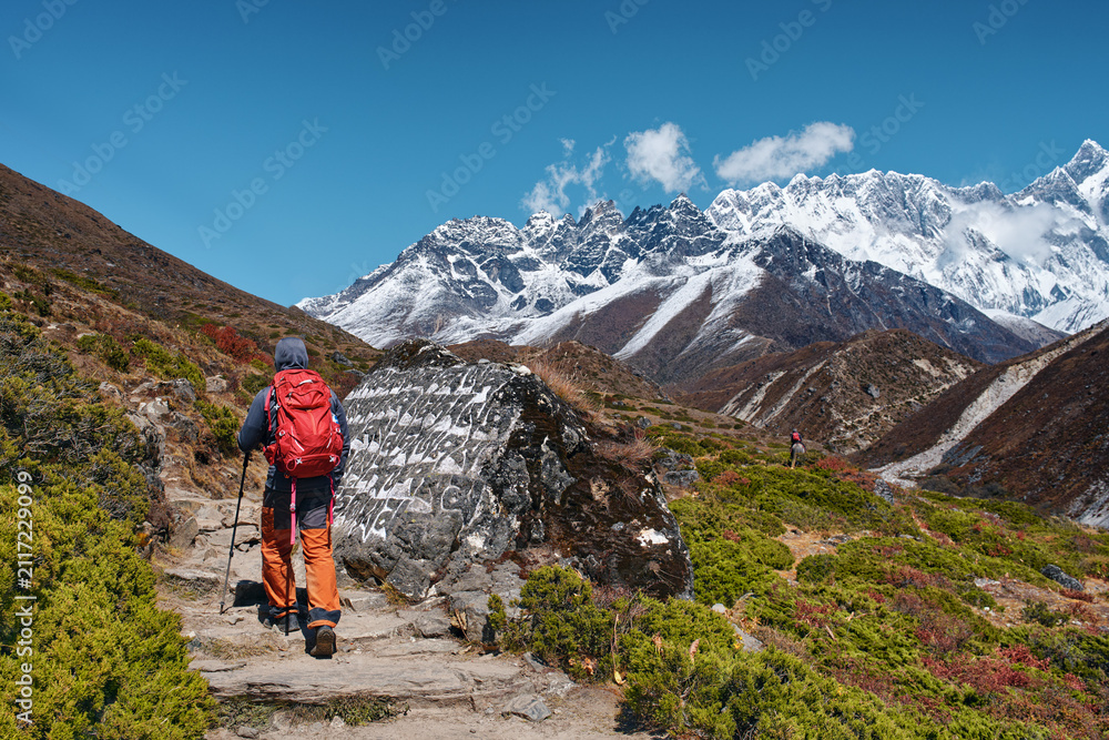 Fototapeta premium Hiker walking on scenic mountain trail past an ancient Buddhist mani stone towards snow-capped peaks in Nepal Himalayas.