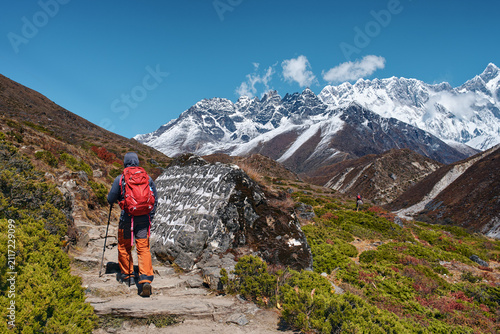 Hiker walking on scenic mountain trail past an ancient Buddhist mani stone towards snow-capped peaks in Nepal Himalayas.