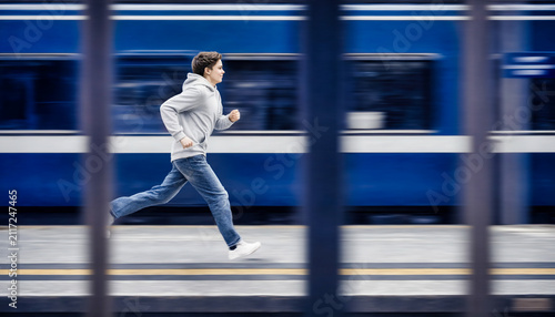 Motion-blurred figure wearing a hooded sweatshirt running past a train platform, viewed through foreground structures, conveying movement, urgency, urban life, and mystery.