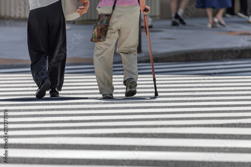 Rear view of an older adult with a walking cane, holding another person's hand as they cross a sunlit urban street.