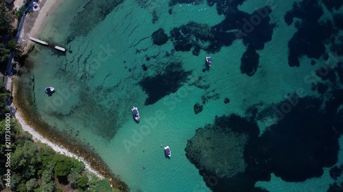 Aerial drone view of a crystal clear turquoise bay on Thassos Island, Greece, with swimmers, paddleboards and a sandy beach surrounded by rocky coastline on a sunny summer day.