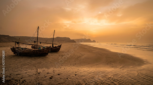 Wooden fishing vessels rest upon a calm sandy shore during a serene golden sunset while gentle ocean waves roll toward the coast under a glowing horizon as daylight fades into the evening calm
