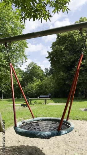 Circular swing with red supports sways gently in a sunny park, surrounded by green grass and trees