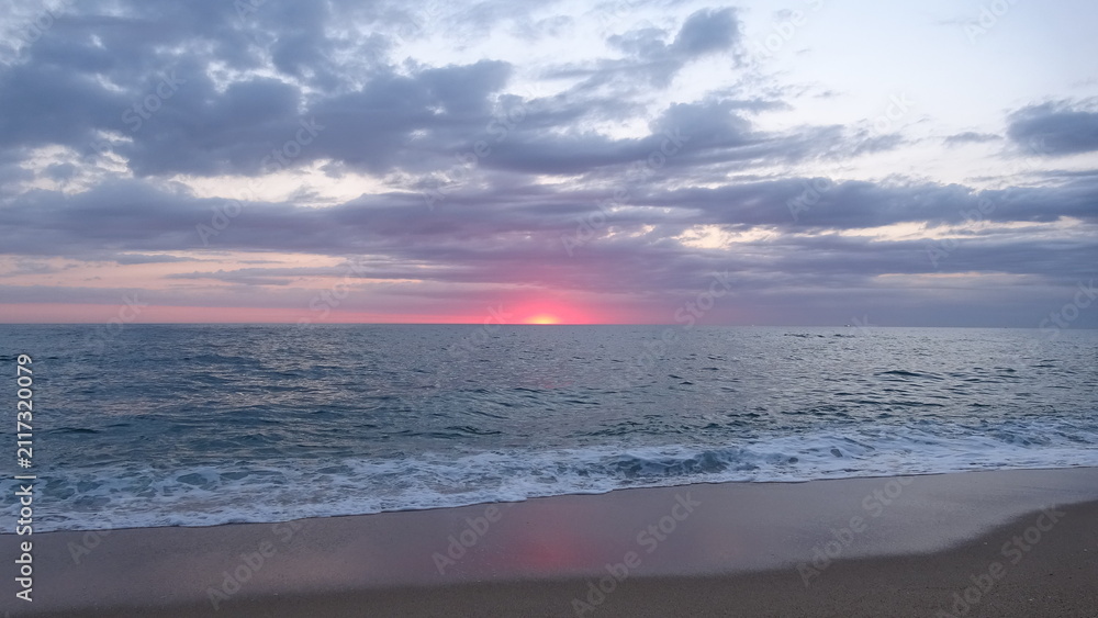 Obraz premium Vibrant Red Dawn Glow on the Sea Horizon. A dramatic red dawn glow lighting up the cloudy sky right before the sun peeks above the ocean horizon on a quiet sand beach.
