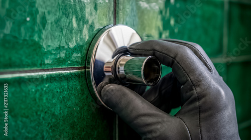 A plumber wearing black gloves attaches a chrome wall flange trim onto a pipe outlet against dark green bathroom tiles.