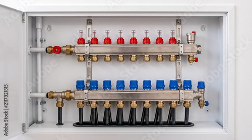 A clean, modern brass heating manifold unit with red and blue control valves mounted on a white utility room wall
