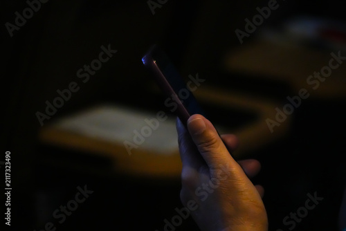 Moody low key silhouette of a hand holding and touching a smartphone screen in a pitch black background.