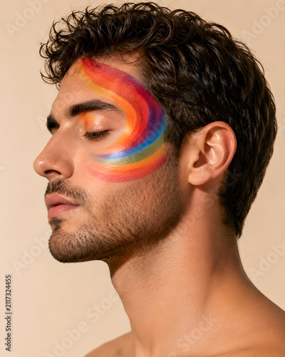 Young man with rainbow face paint posing against beige background  