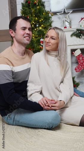 Happy young couple holding hands at Christmas, romantic husband and wife looking at each other seat near decorated Xmas tree and fireplace, cozy winter holiday celebration, love concept
