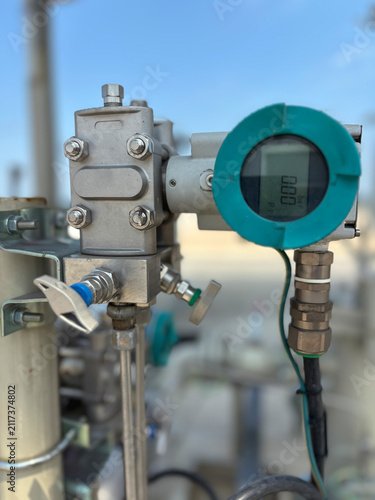 Close-up of a digital pressure transmitter gauge with a turquoise ring and clear screen mounted on industrial piping infrastructure against a clear blue sky.