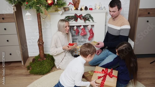 Happy family exchanging Christmas gifts at home, parents with children sharing craft paper presents near decorated fireplace, cozy winter holiday celebration, New Year surprise concept. Vigilia