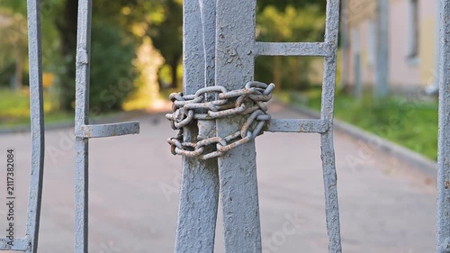 A metal gate is secured with chains, showing a close-up of the lock mechanism. The background features a blurred pathway and greenery, suggesting an outdoor area that is currently inaccessible