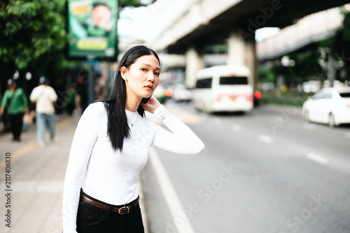 Urban fashion portrait of a young Asian woman standing by a city street in Bangkok, Thailand. wearing a minimalist white long-sleeve top and black trousers with a soft blurred traffic background.
