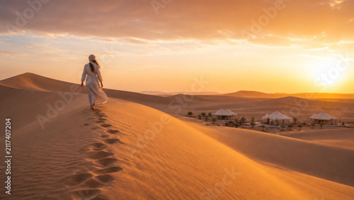 Woman walks alone on golden desert dunes at sunset, leaving footprints behind, with luxury tents visible in distance, evoking calm and solitude
