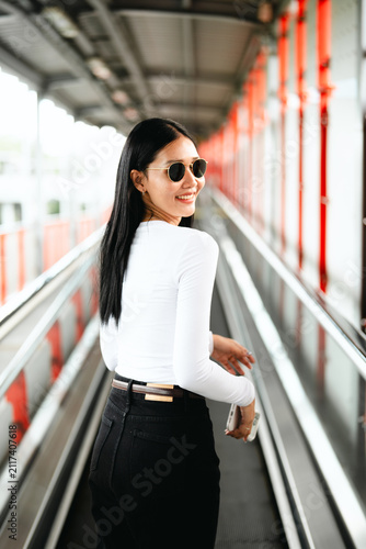 Rear view of a happy Asian woman looking back over her shoulder with a bright smile, wearing a minimalist monochrome outfit and sunglasses while walking on a city skywalk. 
