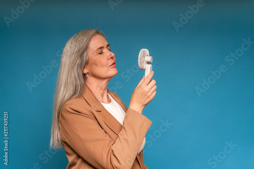 Senior woman cooling down with portable fan against blue background