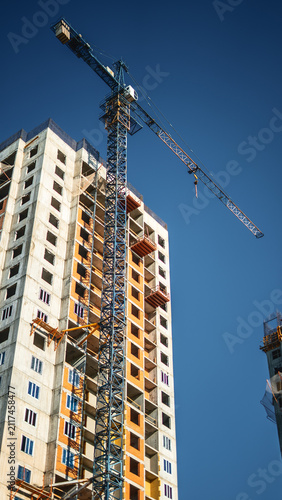 Modern residential tower rises beneath clear blue sky while tower crane supports ongoing construction. Apartment development reflects urban growth, engineering precision and real estate investment.