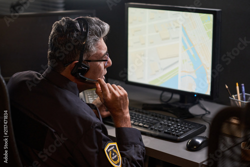 Middle aged security guard monitoring digital map on desktop computer while speaking through headset in control room. Ideal for surveillance, dispatch, safety service marketing