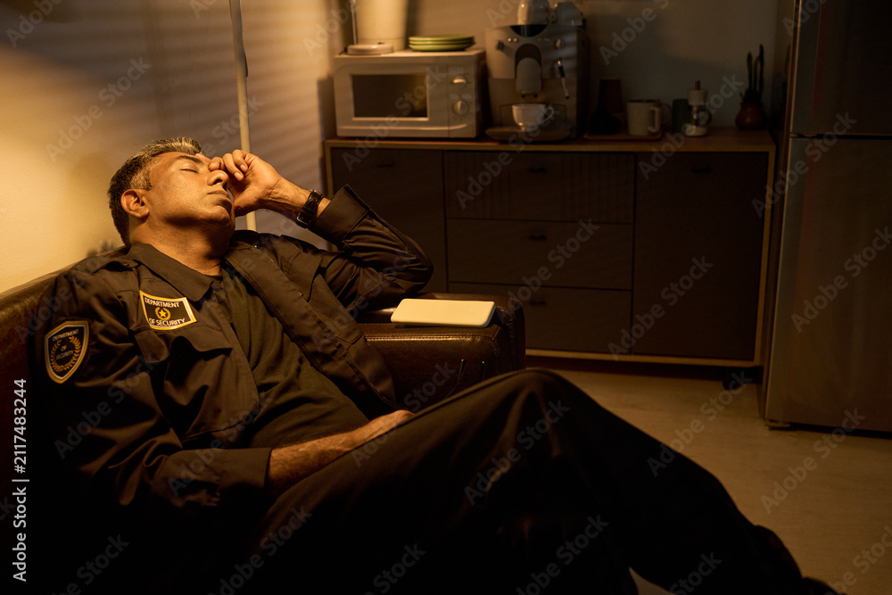 Fototapeta premium Middle aged security guard resting on sofa during break, rubbing forehead in office kitchen, conveying fatigue from long shift. Useful for workplace stress, burnout, night duty campaigns