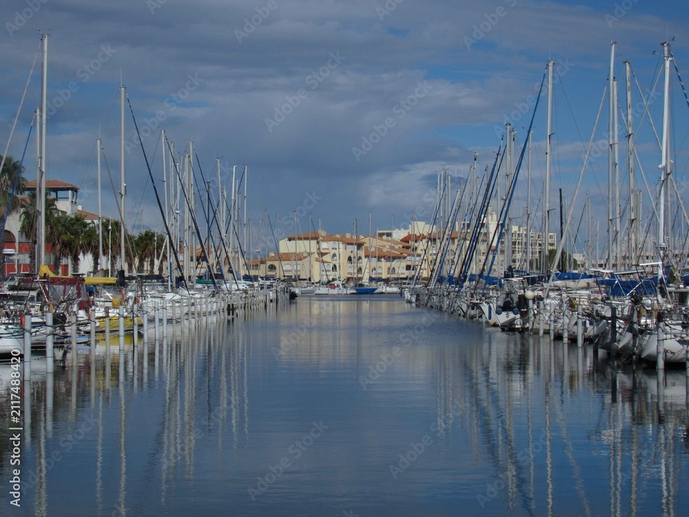 Obraz premium Symmetrical view of sailboats moored in a calm Mediterranean marina with water reflections and waterfront architecture.