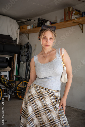 Young woman poses in a garage wearing a gray top and plaid skirt with sunglasses on her head during daytime