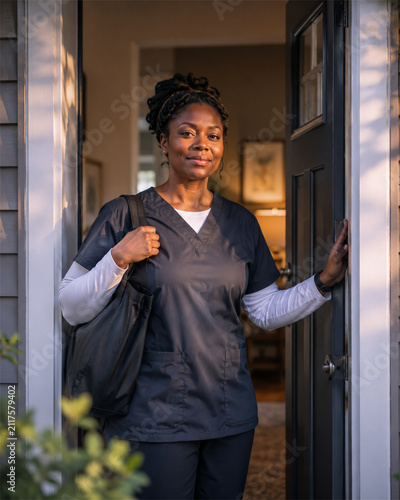 Confident black female healthcare worker wearing medical scrubs and carrying a shoulder bag while standing in a residential doorway ready for a home visit or community care shift in the neighborhood