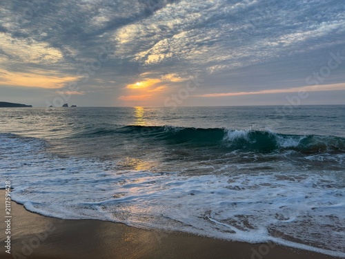 A rising dark emerald wave catches the last rays of sunset over the calm Atlantic. Dramatic textured clouds glow above shimmering reflections, creating a peaceful sunset with natural color and light.