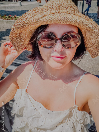 Woman with short dark hair wearing a straw hat and oversized sunglasses poses outdoors, sunlight filtering through the hat, with a cobblestone street in the background