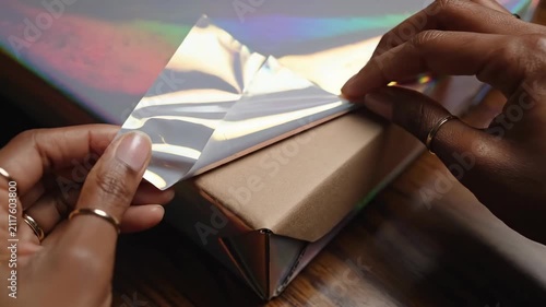 Close-up of hands wrapping a gift box with iridescent holographic paper, preparing for a special occasion.