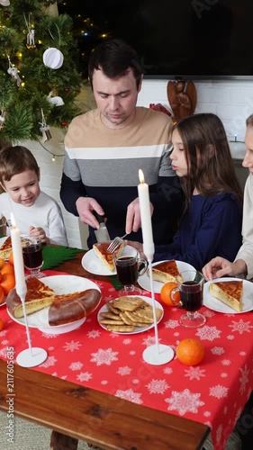 Happy family enjoys Christmas Eve dinner at festive table decorated with red tablecloth, candles, and mandarins. They share traditional holiday cake next to glowing New Year tree. Winter holiday 