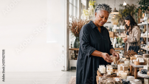 Mature woman arranges handmade candles on wooden displays in a bright boutique with shelving and plants. The composition leaves ample copy space on the left for text or design.