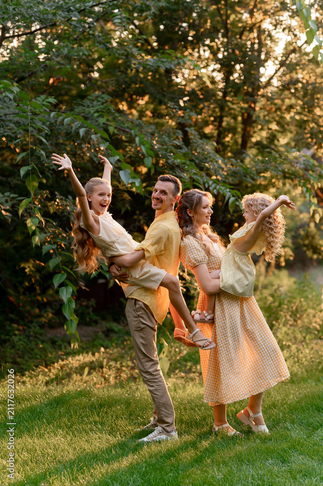 Fototapeta premium A happy family is playing outdoors in the park in summer. Dad, mom, and two daughters are hugging and laughing