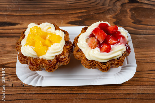 Two flaky puff pastry tarts topped with cream, fresh mango cubes and sliced strawberries on a white paper plate