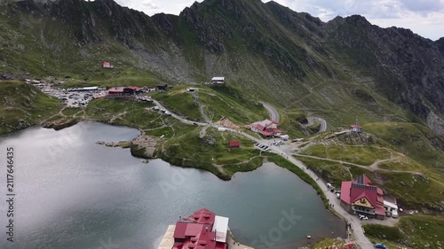 Forward and ascending drone flight revealing Bâlea Lake, mountain restaurants, visitors and parked vehicles surrounded by the dramatic peaks of the Făgăraș Mountains in Romania.