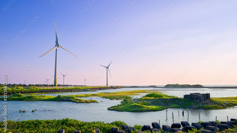 Fototapeta premium Wind turbines stand tall over scenic coastal wetlands with green grass and calm water under a clear sky in Yuanli, Miaoli, Taiwan.