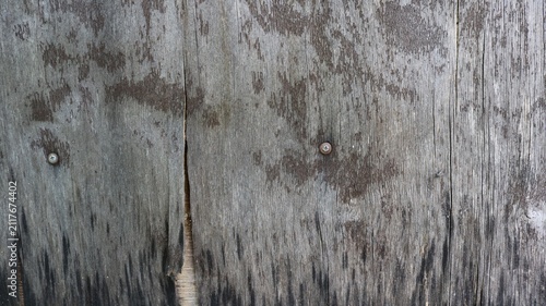 An old gray wooden surface or plywood sheet. The wood grain has dried out, darkened underneath, and is covered with moisture cracks. Two rusty screws are screwed into the surface.