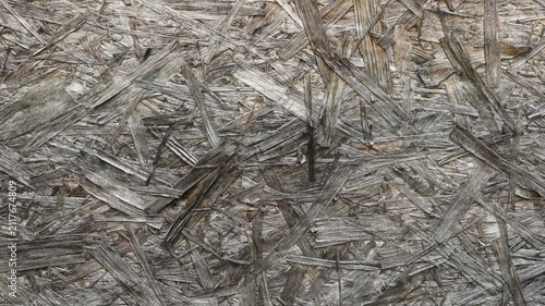 The textured surface of an OSB board made from pressed wood chips. The texture is aged, rough, and chaotic. The wood fibers have faded with age and acquired a noble gray-brown hue.