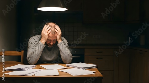 Stressed man sits at kitchen table with paperwork under dim light at night