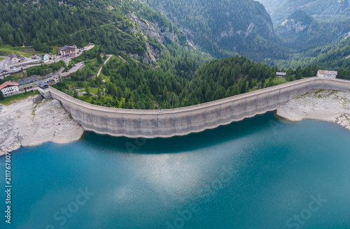 Aerial view of Fedaia Dam and reservoir with low water levels surrounded by the Dolomite mountains and pine forests in Italy.
