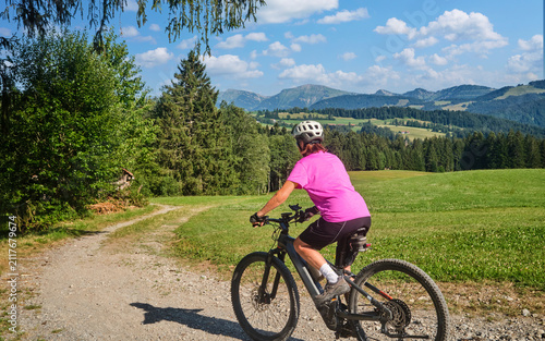 Active senior woman riding an e-mountain bike on a scenic alpine trail beneath the Nagelfluh mountain range on a sunny summer day. Oberstaufen, Allgäu, Bavaria, Germany.