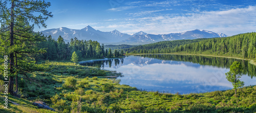 View of a mountain lake on a summer morning, picturesque reflection of the sky with clouds, snow-capped peaks and forested shores