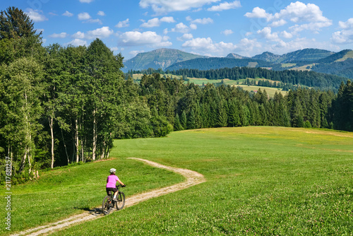 Active senior woman riding an e-mountain bike on a scenic alpine trail beneath the Nagelfluh mountain range on a sunny summer day. Oberstaufen, Allgäu, Bavaria, Germany.