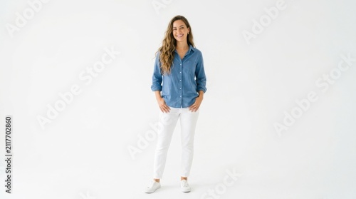 Happy young woman with long brown hair wearing a blue denim shirt and white pants standing and smiling at the camera