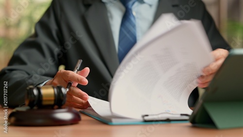 Canvas Print Professional male attorney wearing dark suit reviews legal document paperwork holding pen near wooden gavel clipboard tablet computer sitting desk courtroom office workspace indoor setting