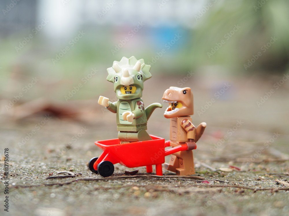 Naklejka premium Indonesia - April 27, 2025 : Mini toy of action figure at outdoor with blurred background. Toy photography concept design. T-rex and triceratops dino characters with trolley wheelbarrow.