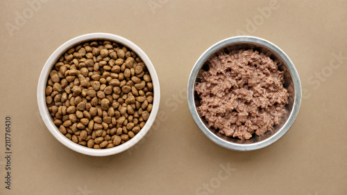 Top view of dry kibble and wet meat in bowls on brown background